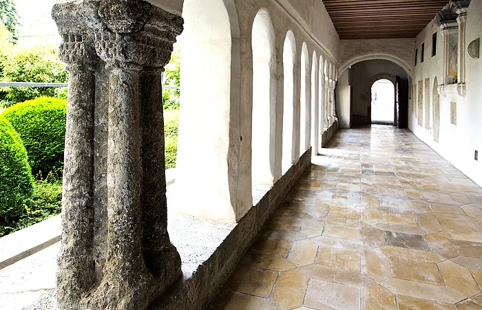 Blick in einen langen Kreuzgang mit Säulen und Rundbögen links im Bild, dahinter grüne Büsche. Rechts im Bild weiße Wand mit einer Ausfräsung in der Wand und Steinplatten mit Text angebracht. Entlang des Gangs alter Bodenbelag aus hellen alten Steinen.