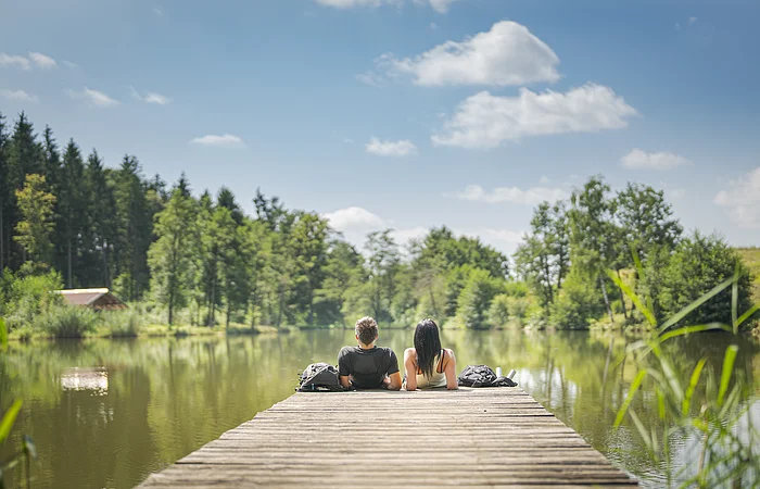 Zwei Personen sitzen auf einem Steg an einem See, umgeben von Bäumen und unter blauem Himmel. Im Hintergrund links im Bild eine kleine Holzhütte zu sehen.