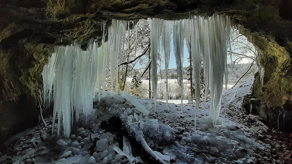 Eishöhle Blick aus einer Höhle mit großen Eiszapfen an der Decke auf eine verschneite Winterlandschaft mit Bäumen.