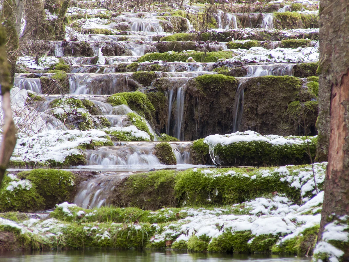 kleine Wasserfälle fließen über moosbedeckte Steinstufen mit leichtem Schneebelag im Wald