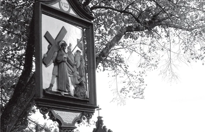Schwarz-weiß Foto der Kreuzweg-Station am Kalvarienberg in Wittesheim mit einem Wegkreuz mit einer Kreuzigungsszene und dem Dorf mit Kirche im Hintergrund, unter Baumzweigen bei Tageslicht.