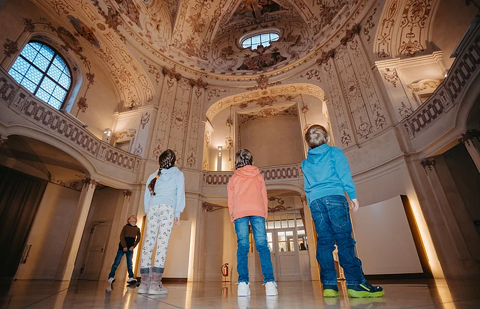 Vier Kinder stehen in einem historischen Raum und schauen zur verzierten Decke mit Fenstern und Wandmalereien.