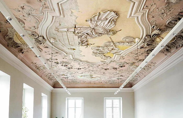 Farbig gestaltete Decke mit barocker Stuck- und Freskoverzierung in einem hellen Raum mit weißen Wände, vielen Fenstern und moderner an der Decke hängender Beleuchtung.