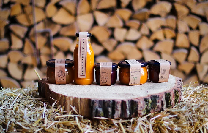 Fünf Gläser mit Kürbisketchup und Saucen auf einem Baumstamm vor gestapeltem Brennholz und Stroh.