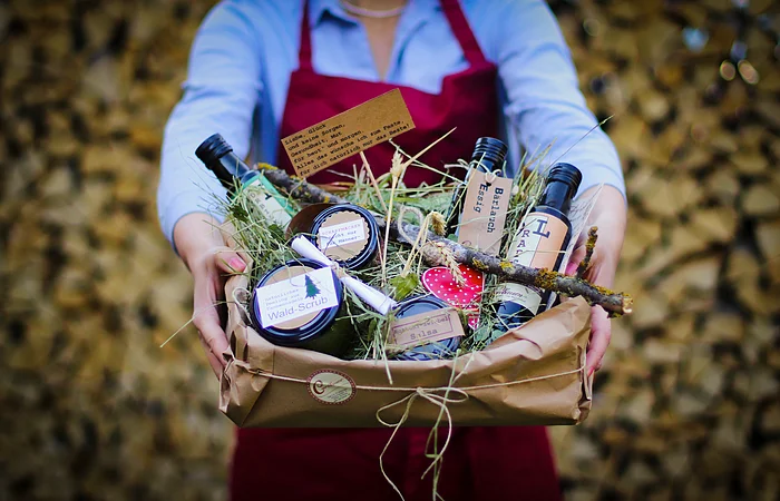 Person in roter Schürze hält Geschenkbox mit Flaschen, Gläsern und Naturdekoration aus Gras und Zweigen.