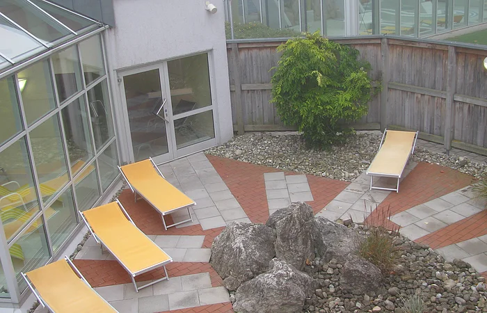 Innenhof mit drei gelben Liegestühlen, großen Steinen und einem Baum, umgeben von Glasfenstern und Holzzaun.