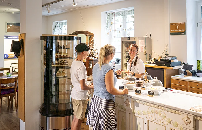 Zwei Leute an Tresen zwischen Kuchenvitrine und Gebäck erhalten Eintrittskarten von Frau