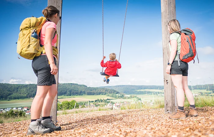 Zwei Erwachsene mit Rucksäcken stehen neben einem Kind auf einer Schaukel, Landschaft im Hintergrund.