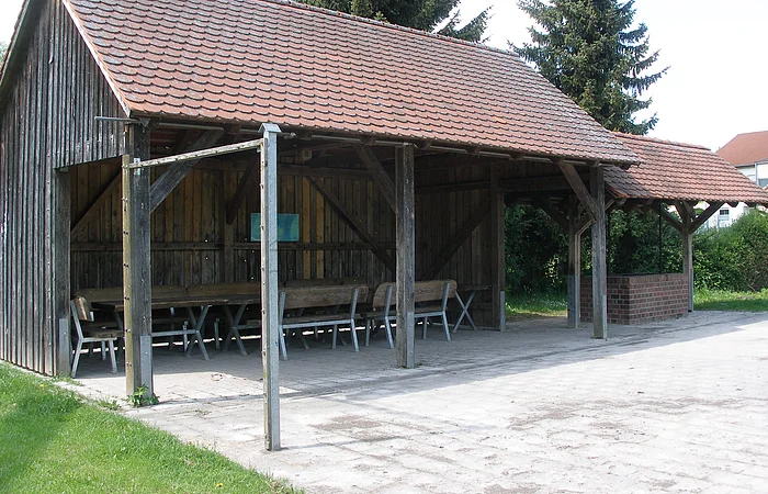 Überdachter Holzpavillon mit Tischen und Bänken auf gepflastertem Boden neben Rasenfläche und Bäumen.