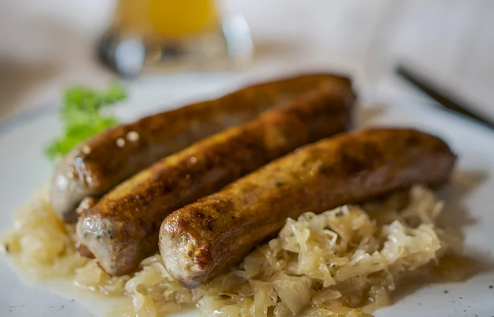 Drei Bratwürste auf Sauerkraut auf einem Teller, im Hintergrund ein Weizenglas mit Bier gefüllt.