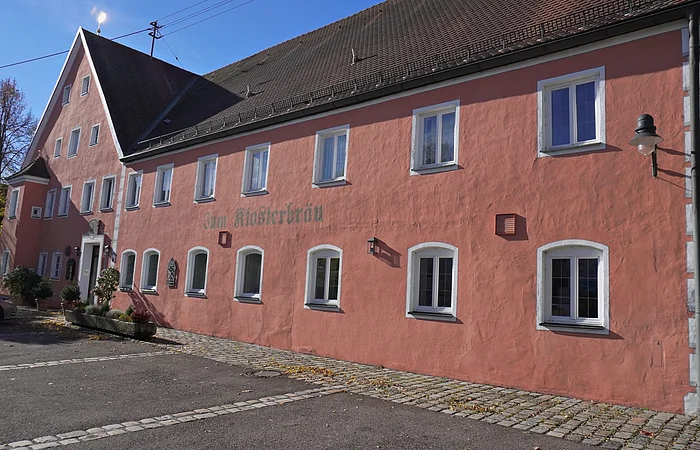 Rosa gestrichenes Gebäude mit weiß gerahmten Fenstern und Pflastersteinen davor bei Sonnenschein.