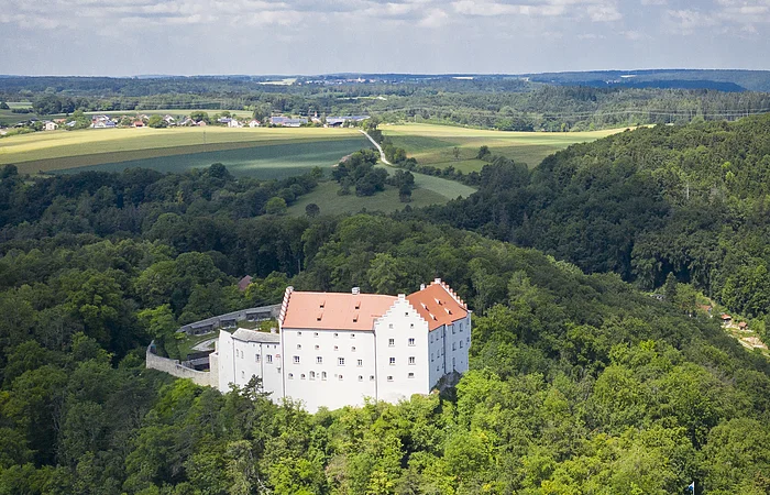 Luftaufnahme eines weißen Schlosses mit rotem Dach, umgeben von Wald und Feldern unter blauem Himmel.