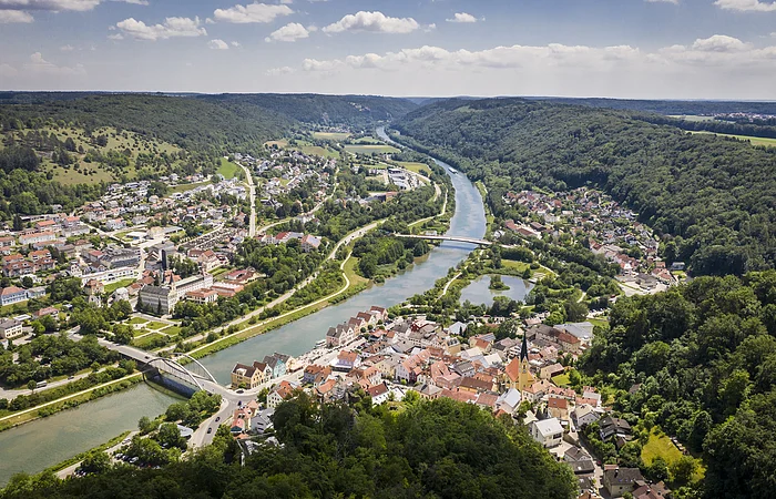 Der Blick auf Riedenburg von oben. Mitten durch die Stadt verläuft der Main-Donau-Kanal. Umrandet wird sie von grünen Wäldern. Der Himmel ist blau und einige Wolken sind zu sehen.