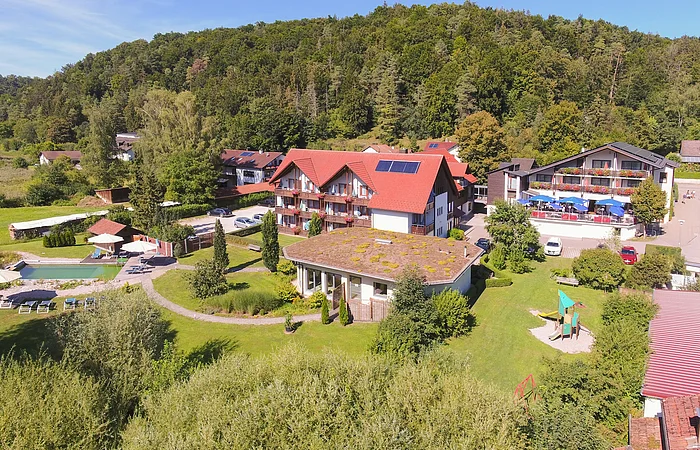 Das Hotel Zur Linde in Schambach von außen. Im Hintergrund Wohnhäuser und ein Wald und im Vordergrund das Hotel mit Außenanlage, Wiese, Pool und vieles mehr.