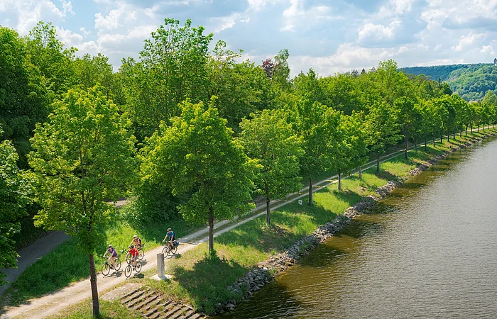 Fünf Radfahrer auf einem Weg entlang eines Flusses mit Bäumen und Hügeln im Hintergrund bei bewölktem Himmel.