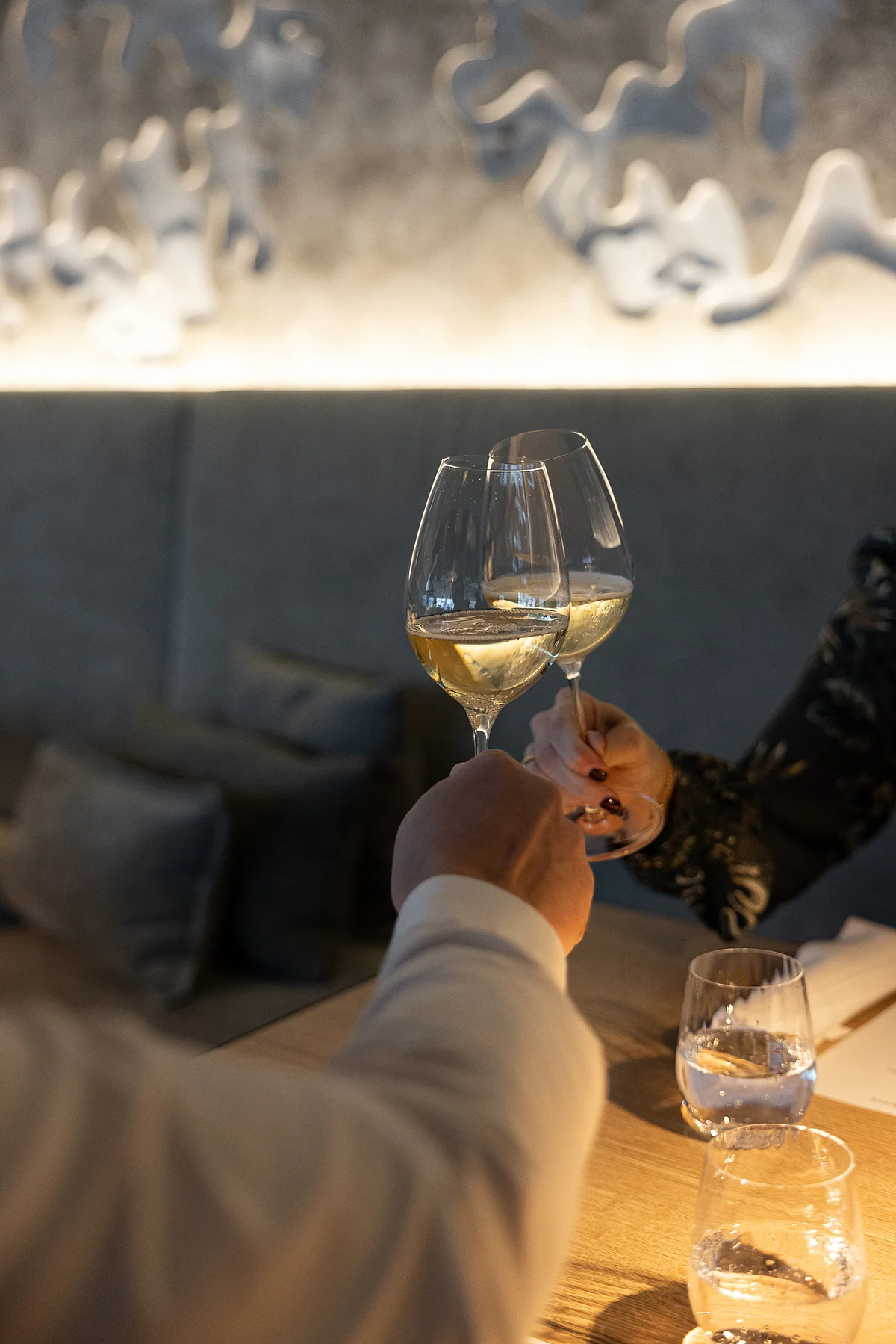 Das Altmühltal – Wo Bollinger pricklet und Genussmomente leuchten Zwei Personen stoßen mit Weißweingläsern an einem Holztisch in einem Restaurant an.