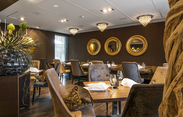 Restaurant mit gedeckten Tischen, gepolsterten Stühlen, drei runden Spiegeln an der Wand und Kronleuchtern an der Decke