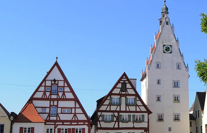 Historische Fachwerkhäuser, ein Torturm mit Uhr in einer Altstadt bei klarem Himmel. Links im Bild parkende Autos auf einem gepflasterten Platz. Rechts im Vordergrund ein Baum mit einer Info-Tafel und einem großen Buchstaben "O" aus Stein. Dahinter eine Informationsvetrine.