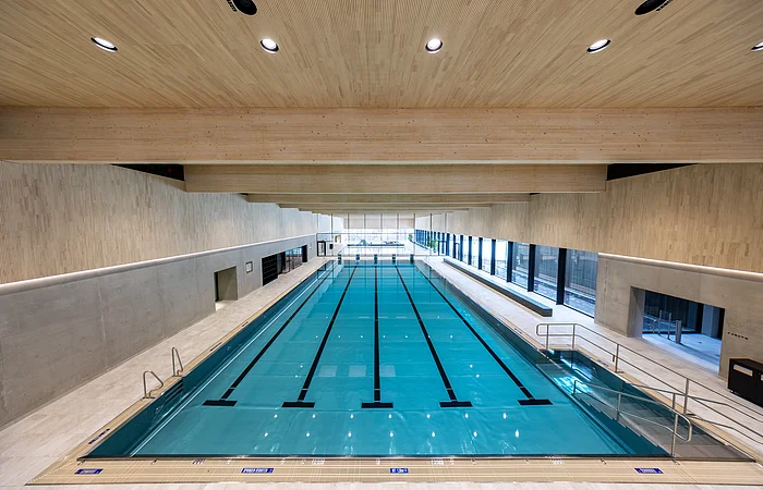 Innenaufnahme eines Schwimmbeckens mit Holzdecke und großen Fenstern an der rechten Seite