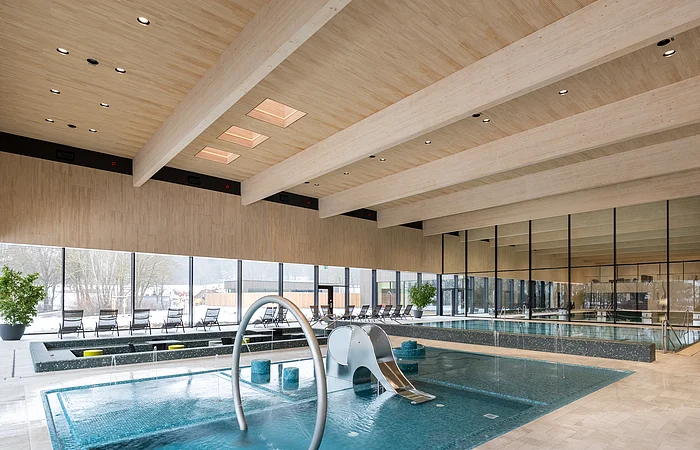 Innenraum eines Schwimmbads mit Kinderbecken, Wasserrutsche und großen Fenstern zur Außensicht.