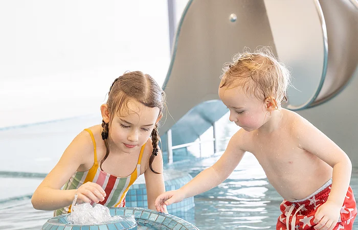 Zwei Kinder spielen in einem flachen Schwimmbecken an einem blauen Mosaikbrunnen mit Wasserspielzeug.
