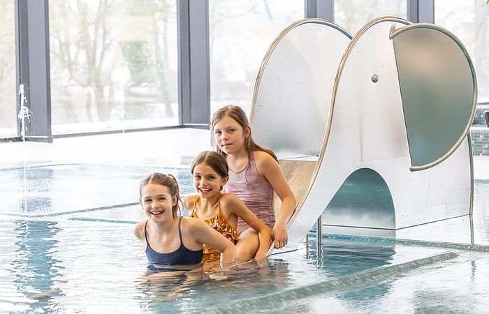 Drei Mädchen in Badebekleidung sitzen an einem kleinen Elefant-Wasserrutschenbecken in einem Hallenbad.