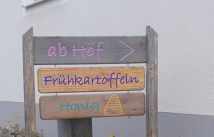 Holzschild mit den Aufschriften „ab Hof >“, „Frühkartoffeln“ und „Honig“ vor Hauswand und Pflanzen.