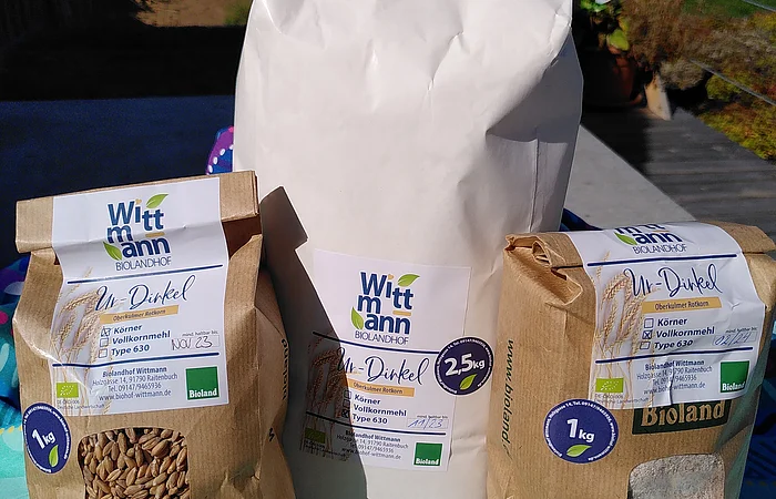 Drei Packungen Ur-Dinkel Vollkornmehl und Körner von Biolandhof Wittmann auf buntem Tuch im Freien.