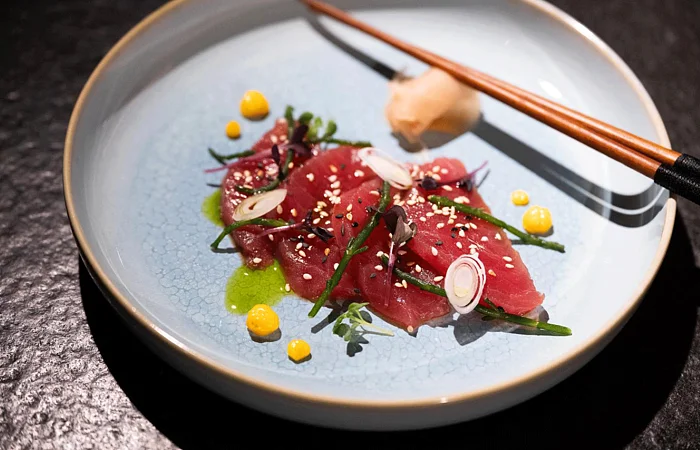 Dünne Scheiben rohen Fischs mit grünen Kräutern, Sesam, gelbem Mais und roten Zwiebeln auf einem schwarzen Tisch im blauen Teller mit Essstäbchen serviert.