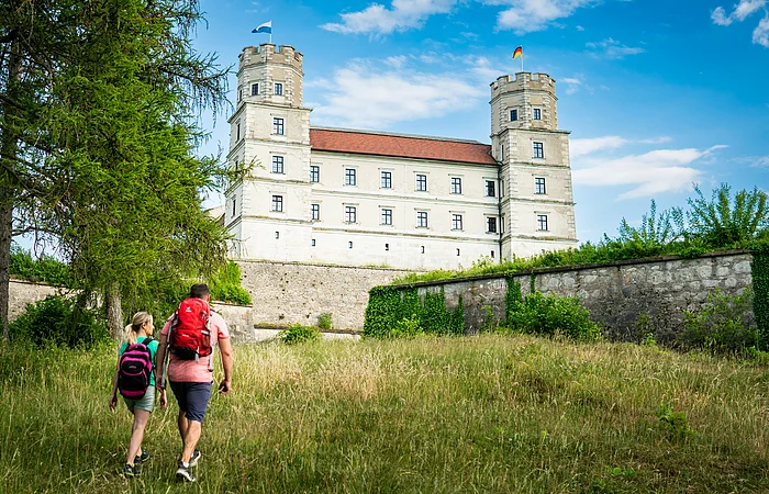 Eine Frau und ein Mann laufen leicht bergauf auf die Fassade einer Burg mit zwei Türmen zu. Sie tragen kurze Hosen, T-Shirts und je einen Rucksack.