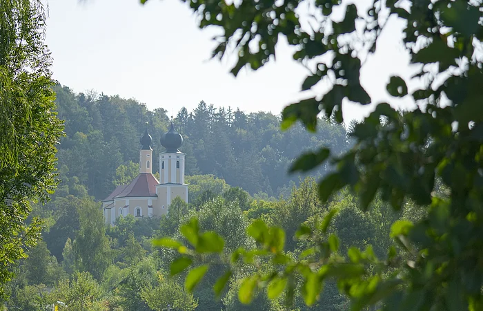 Ein Pärchen wandert unterhalb der Wallfahrtskirche Breitenbrunn entlang satter grüner Wiesen. Die Wallfahrtskirche ist von grünen Wäldern umgeben.