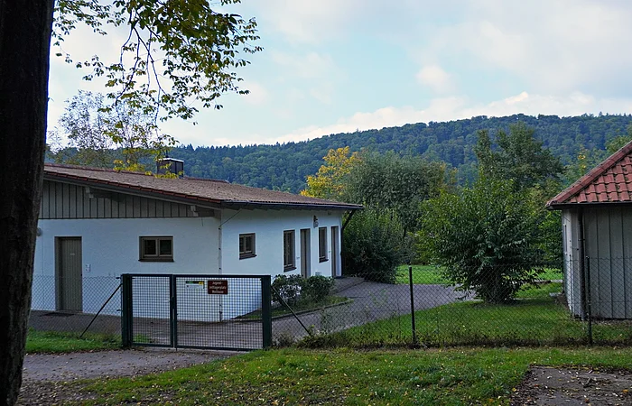 Eingezäuntes Grundstück mit zwei Gebäuden, Bäumen und bewaldetem Hügel im Hintergrund bei Tageslicht.