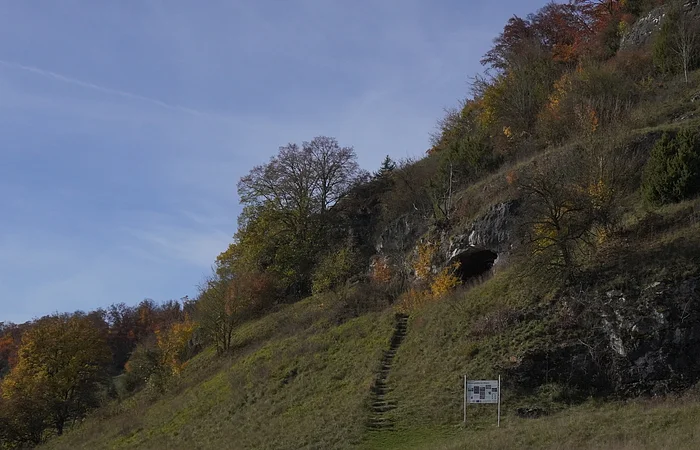 Grasbewachsener Hang mit Treppe, Bäumen und einer Felsöffnung unter blauem Himmel.