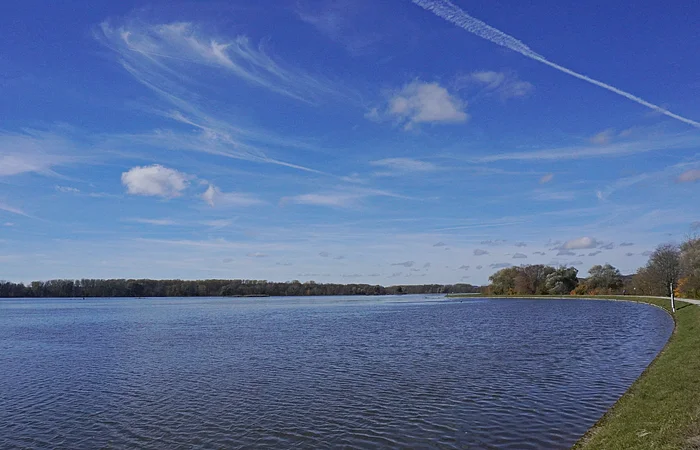 Uferweg entlang eines großen Gewässers unter blauem Himmel mit wenigen Wolken und Baumreihe am Horizont.