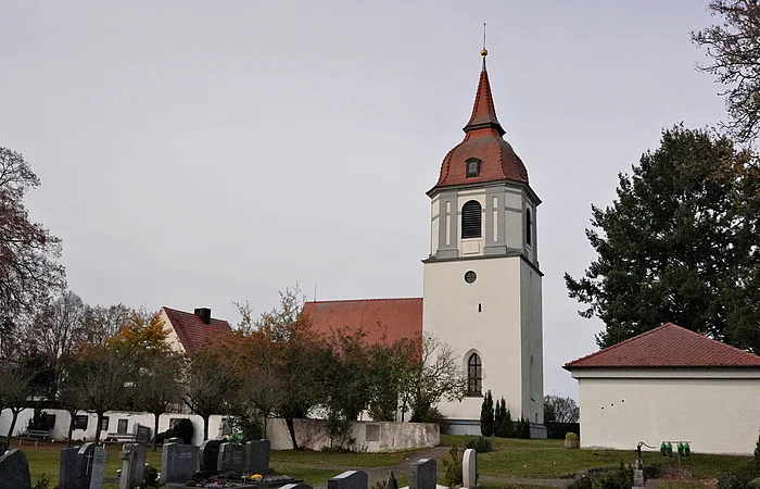Kirche mit Turm und rotem Dach in einem Friedhof mit Grabsteinen und Bäumen bei bewölktem Himmel.