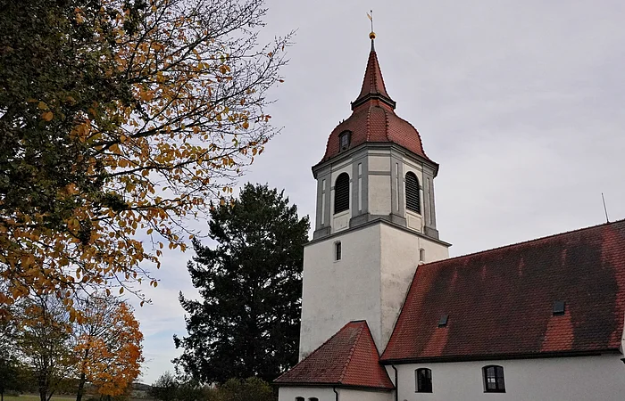 Kirchturm mit rotem Dach neben Bäumen mit herbstlichen Blättern unter bewölktem Himmel.