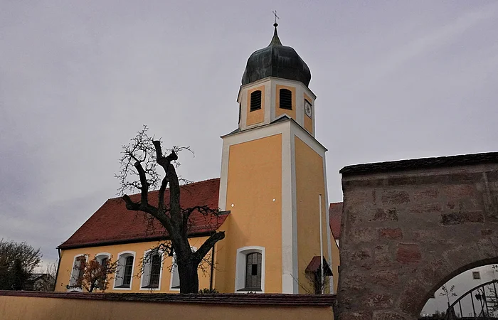 Kirchturm mit Zwiebeldach und gelbes Kirchengebäude hinter einer Mauer bei bewölktem Himmel.