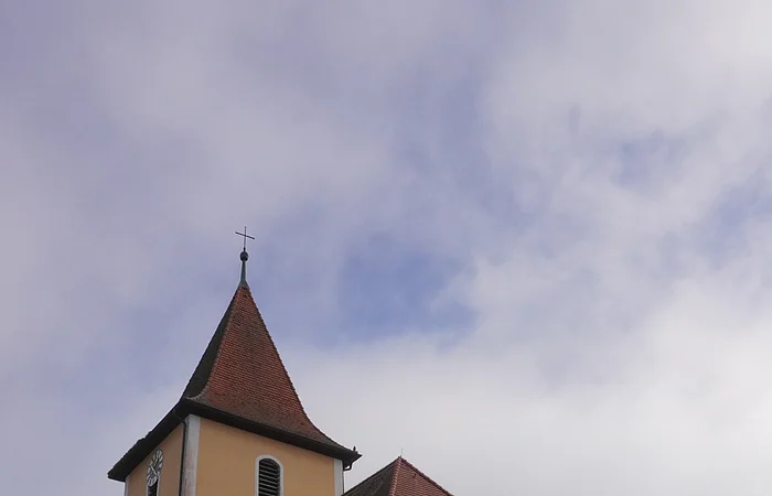 Kirche mit gelber Fassade, rotem Dach und Turm mit Kreuz vor bewölktem Himmel.