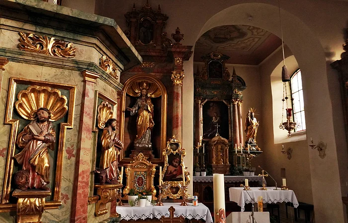 Innenansicht einer Kirche mit Altären, goldverzierten Heiligenfiguren und Kerzen auf Tischen mit weißen Tüchern