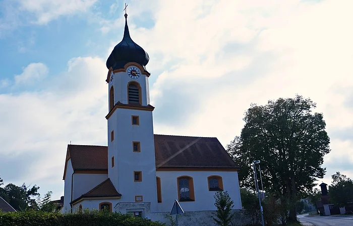 Kirchturm mit Zwiebelhaube und Uhr an weißem Kirchengebäude bei bewölktem Himmel und Bäumen im Hintergrund.