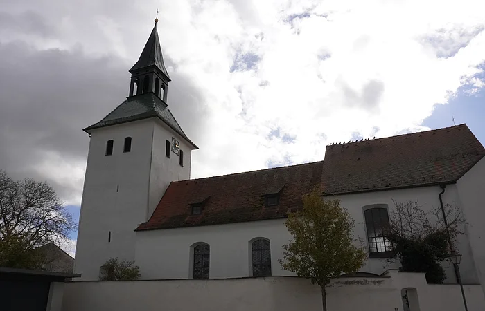 Kirche mit Turm und Uhr vor bewölktem Himmel, Bäume und Mauer im Vordergrund.