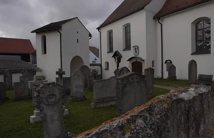 Friedhof mit alten Grabsteinen vor weißen Kirchengebäuden unter bewölktem Himmel.