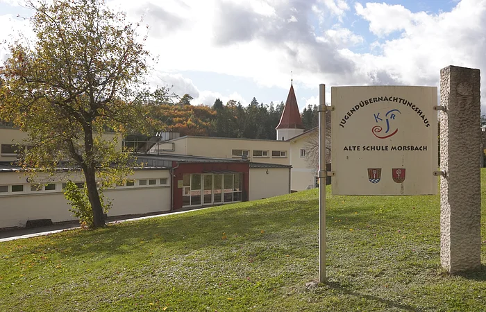 Schild mit Aufschrift „Jugendübernachtungshaus Alte Schule Morsbach“ vor einem Gebäude und Baum auf einer Wiese