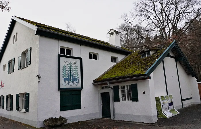 Weißes zweistöckiges Haus mit grünem Fensterladen und Moos auf dem Dach, im Herbstwald gelegen.