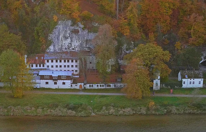 Gebäude vor bewaldetem Felsen mit herbstlich gefärbten Bäumen an einem Flussufer.
