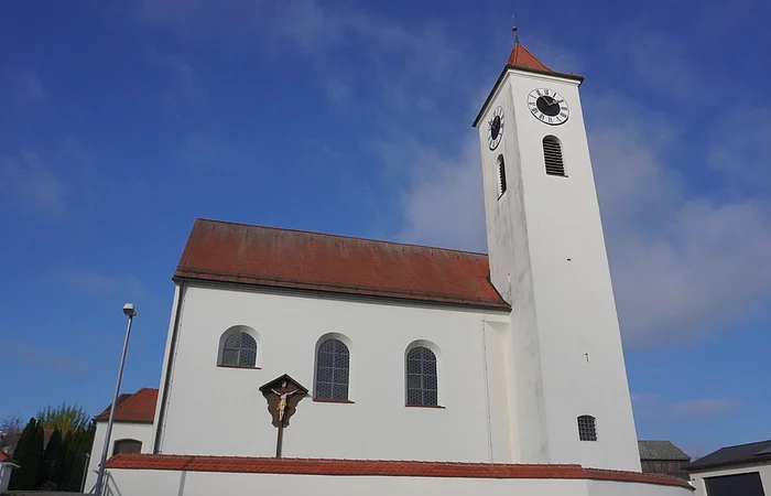 Kirchturm mit Uhr und weißes Kirchengebäude unter blauem Himmel, Vorderseite mit drei Fenstern und Kreuz am Gebäude.