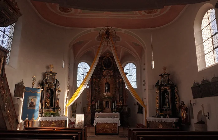 Innenraum einer Kirche mit Altar, zwei Fenstern und hängenden gelben Stoffbahnen am Altarbereich.