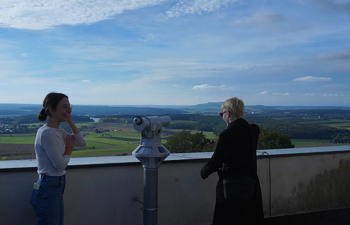 Zwei Personen stehen an einer Aussichtsplattform mit Fernrohr und Blick auf eine weite Landschaft mit Feldern und Wald.