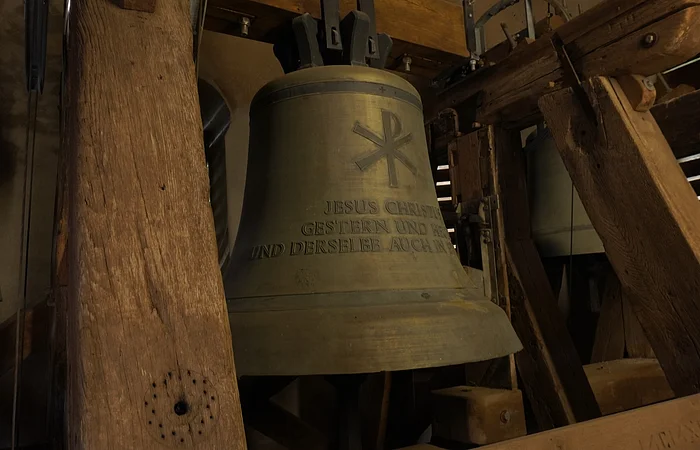 Große bronzene Kirchenglocke mit christlichem Symbol und Schriftzug, umgeben von Holzbalken im Glockenturm.