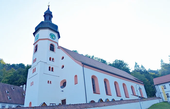 Kirchturm mit Uhr an weißem Kirchengebäude vor bewaldetem Hügel unter hellem Himmel